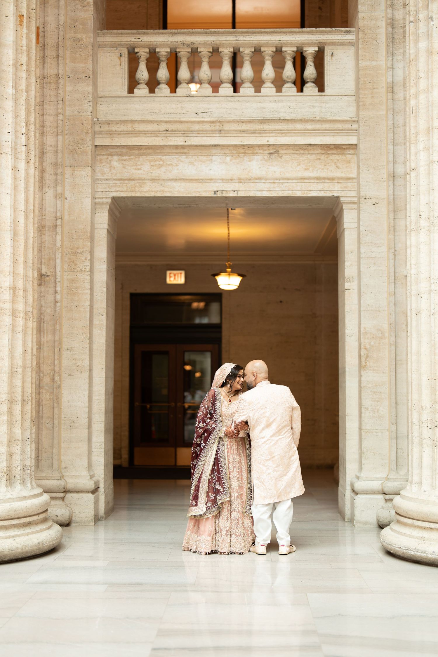 South Asian bride and groom share an intimate moment framed by marble columns in a grand hall.