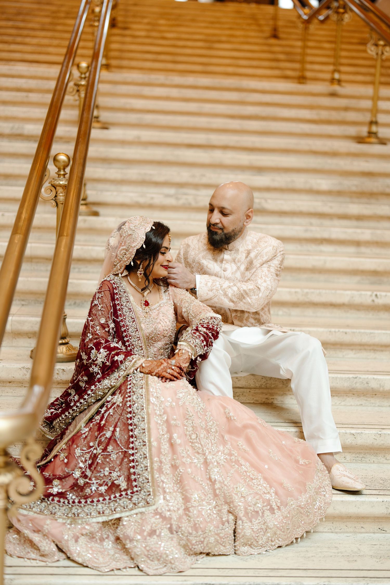 South Asian couple in traditional wedding attire sit together on grand marble staircase with gold railings.