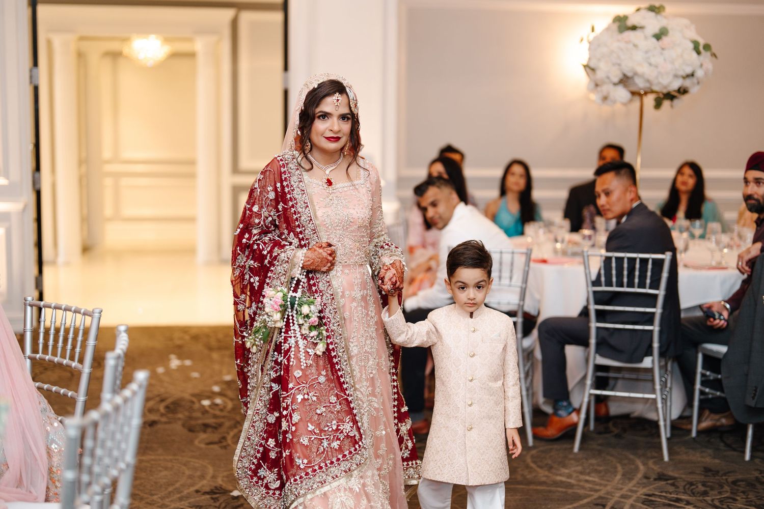 South Asian bride in red and pink bridal lehenga walks hand-in-hand with young boy at wedding reception.