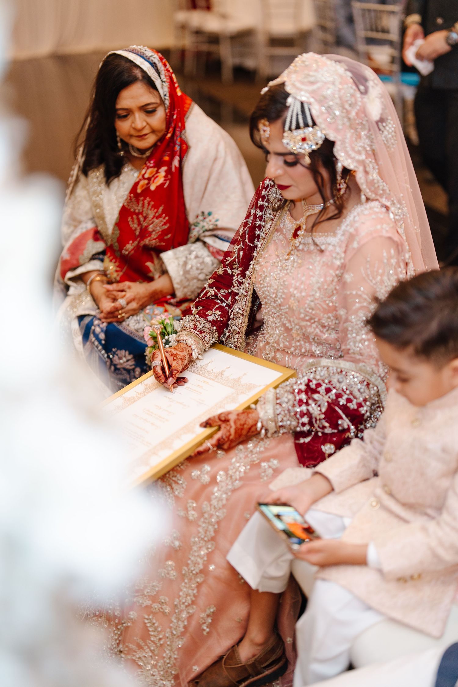 Pakistani bride signing nikah papers in pink embroidered bridal dress with guests during wedding ceremony.