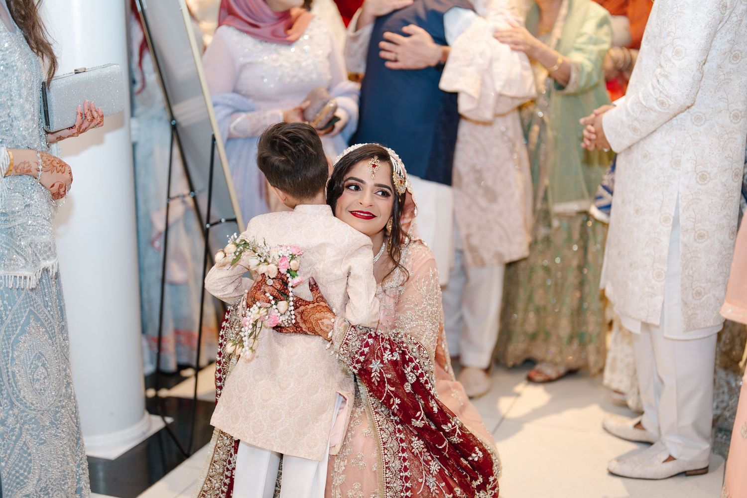South Asian bride in red lehenga shares a warm hug with a young boy in cream sherwani.