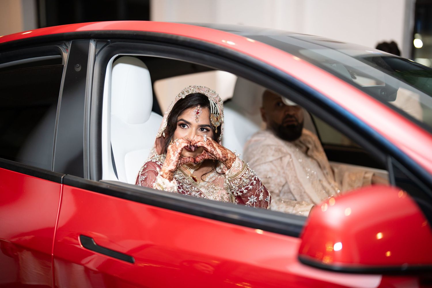 South Asian bride in red bridal attire poses inside a red car showing her henna-adorned hands.