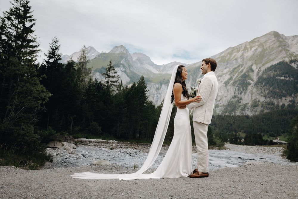 A couple in wedding attire embrace against a backdrop of majestic mountain peaks and evergreen forest in a dreamy outdoor setting.