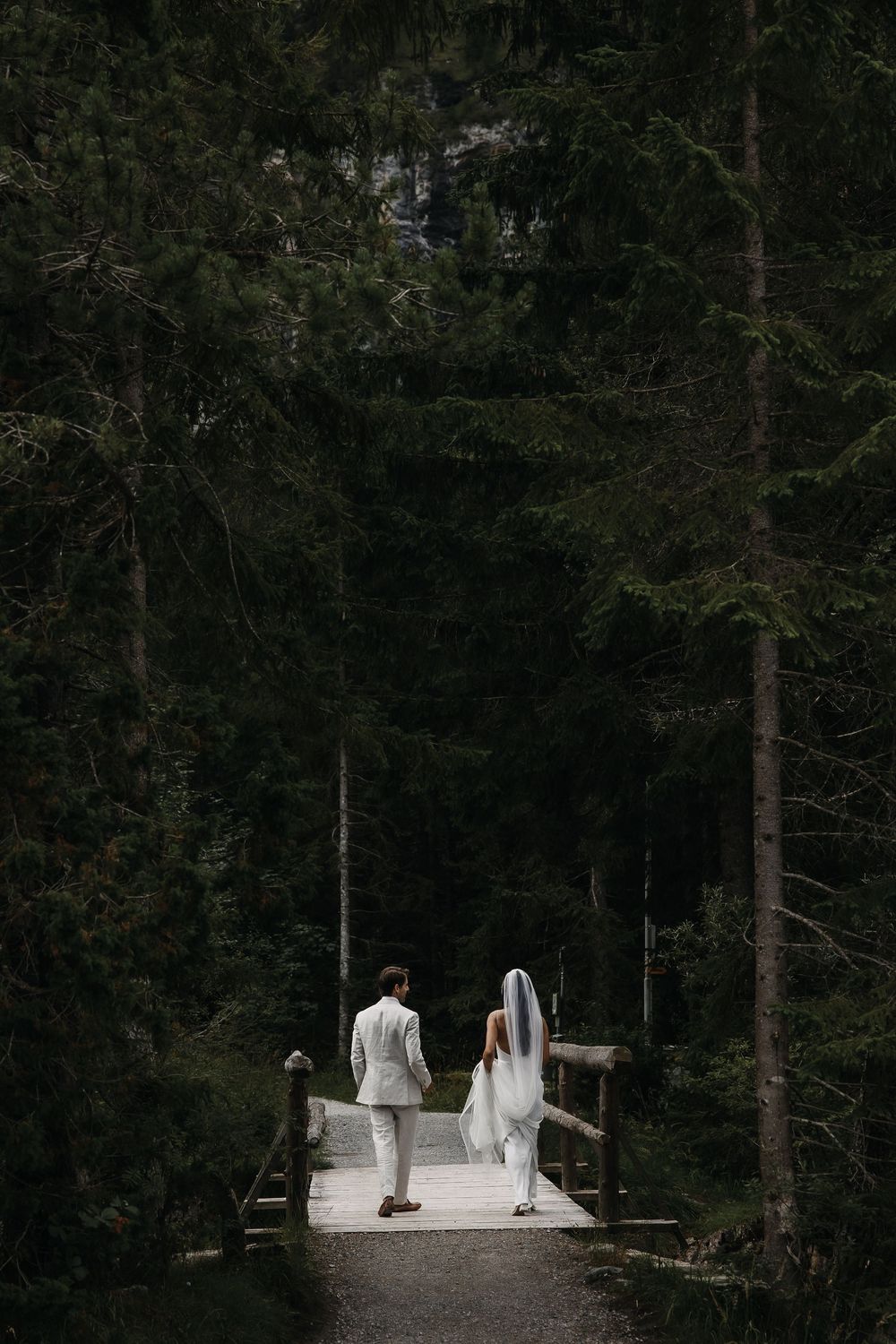 A person in a flowing white dress walks across a wooden bridge in a dark forest setting.