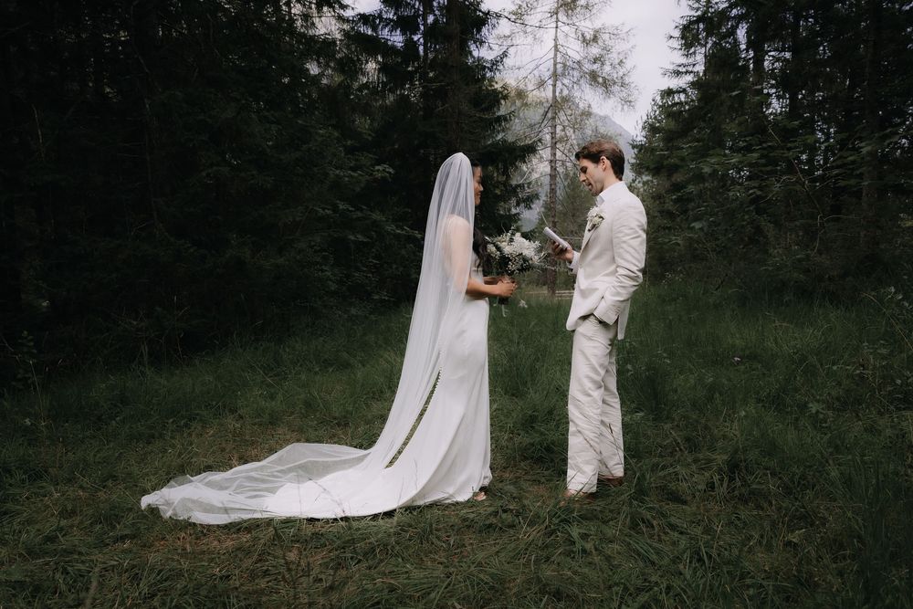 A couple in wedding attire stands outdoors for rustic first look photos in a wooded setting.