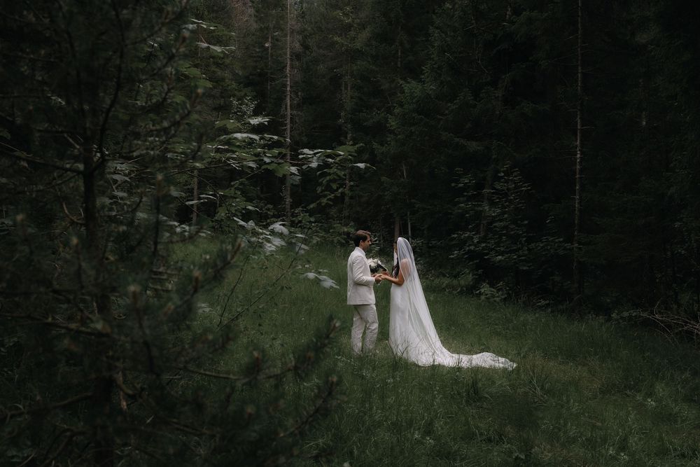 Newlyweds walk through a lush forest path wearing formal wedding attire as sunlight filters through the trees.