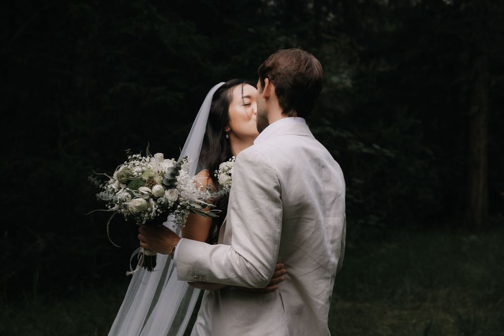 Newlyweds share intimate moments during outdoor wedding photoshoot against dark forest background.