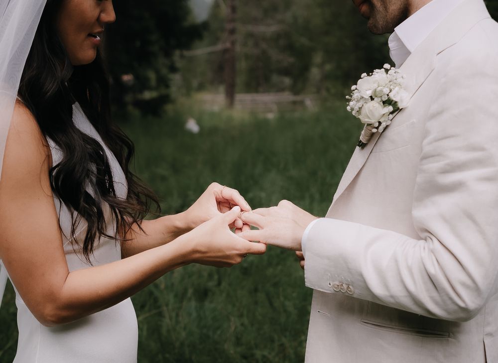 Two people hold hands during an outdoor wedding ceremony with white bouquets against a lush green background.