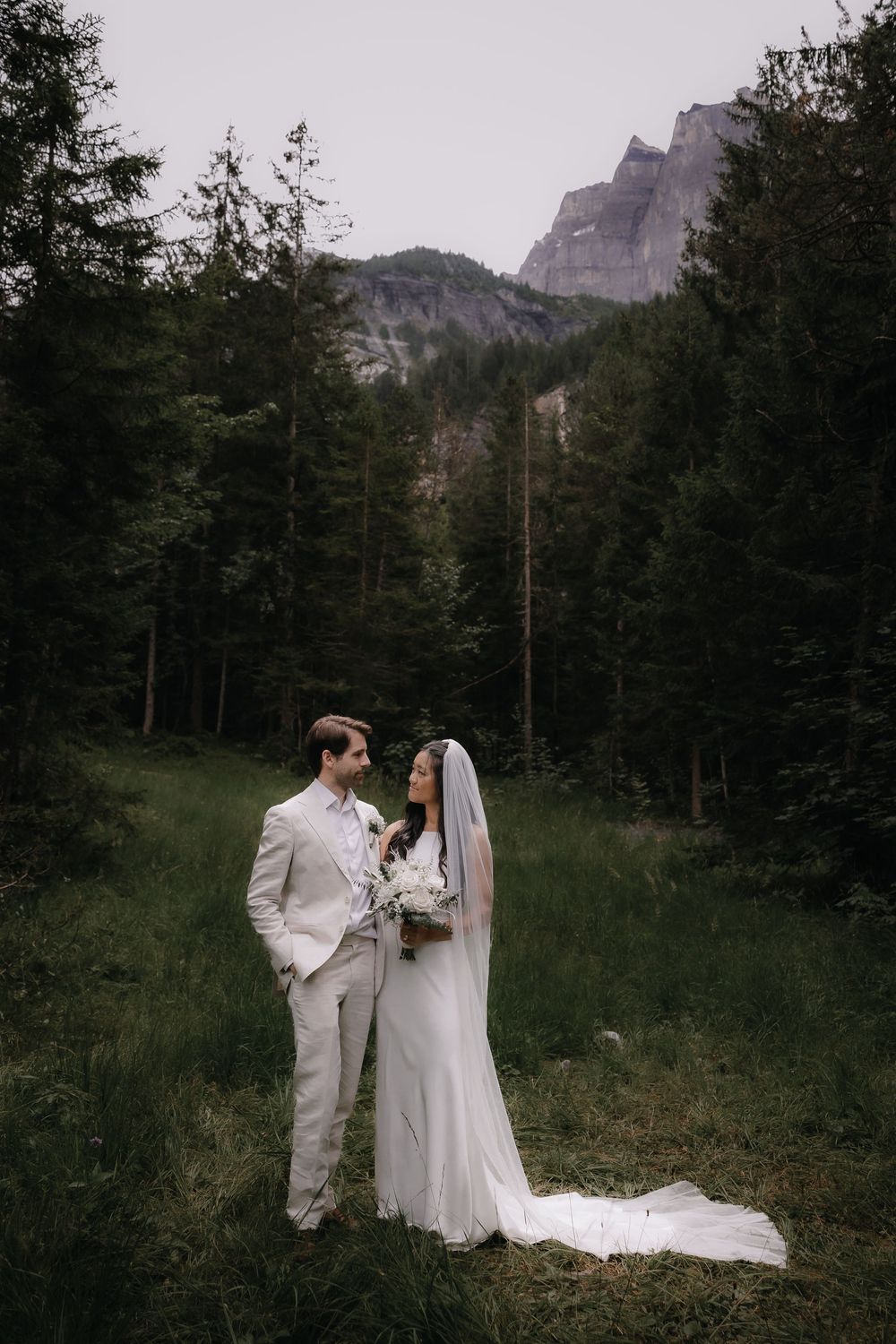 A wedding couple shares an intimate moment in a mountain forest clearing beneath towering peaks.