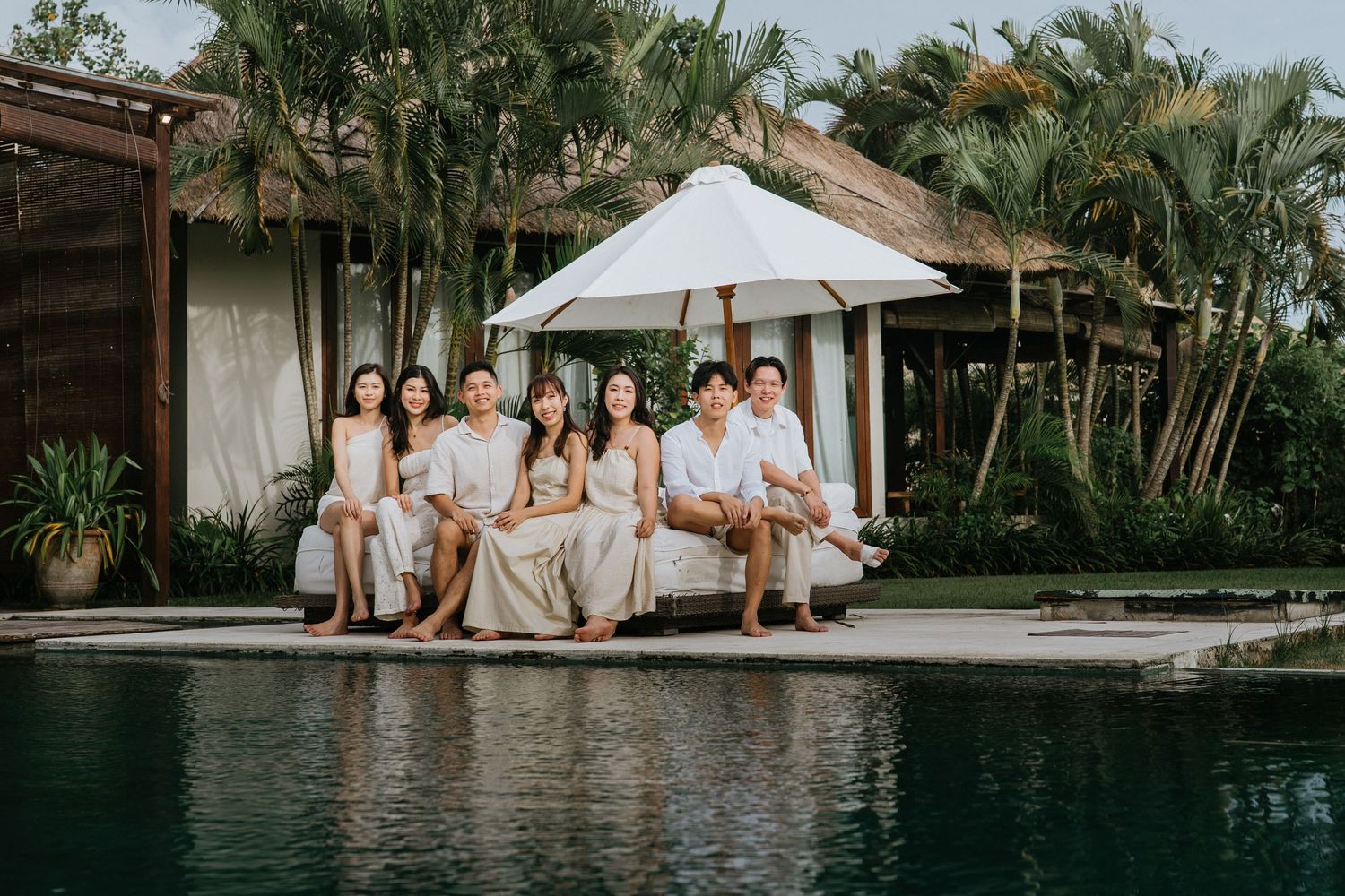 A group of people in white outfits sit together by a luxurious pool surrounded by tropical palm trees.