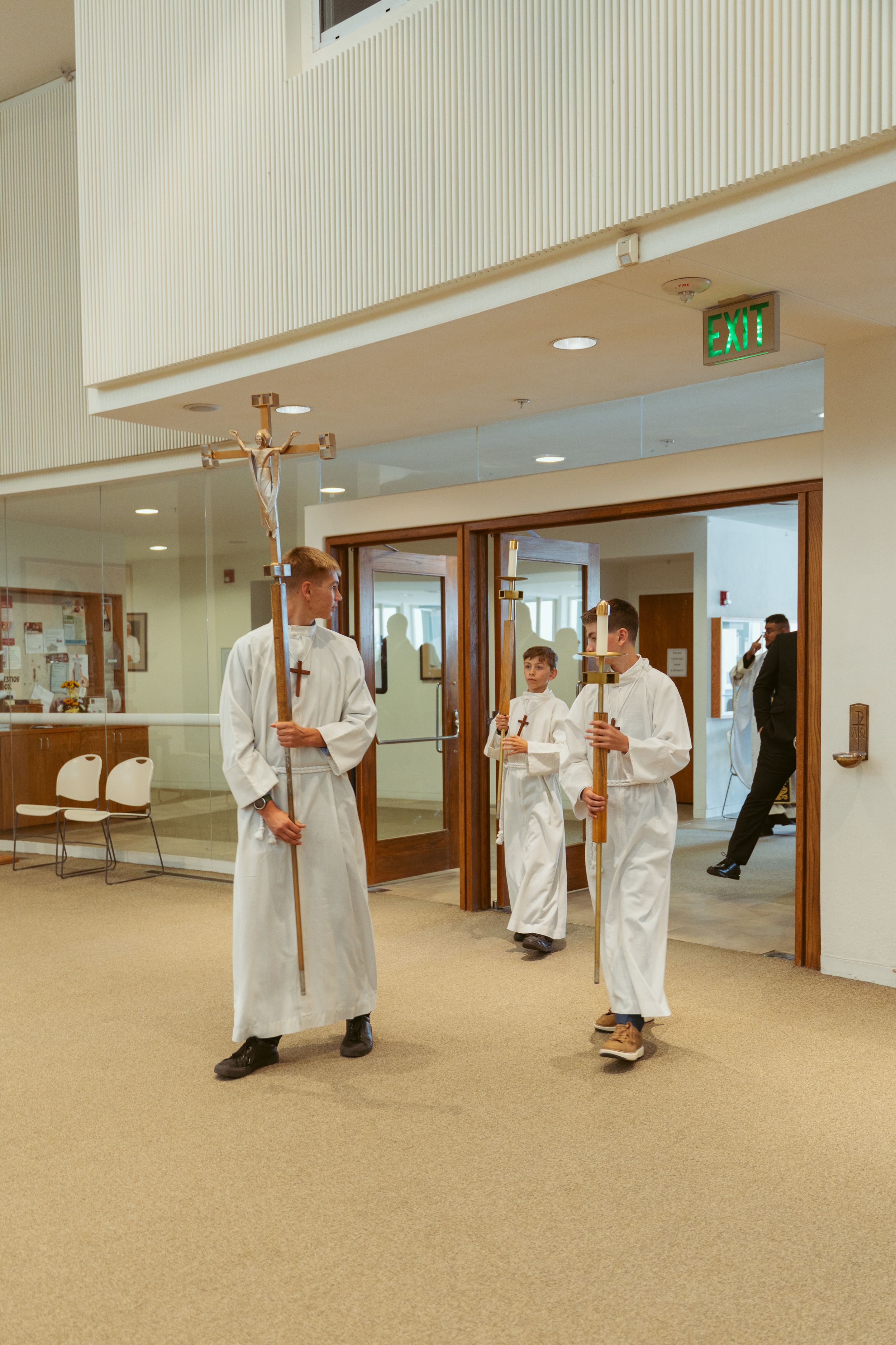 Three altar servers in white robes stand in a church hallway near wooden doors and a green exit sign.