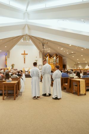 Three altar servers stand at the front of a modern Catholic church interior with wooden pews and organ pipes.
