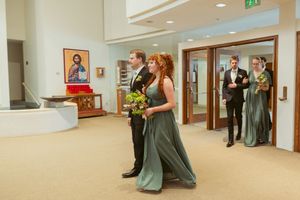 Wedding party members walk through a modern hallway with sage green dresses and floral bouquets.