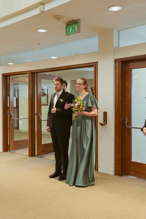 A couple stands ready in formal attire in a hallway before entering through wooden doors.