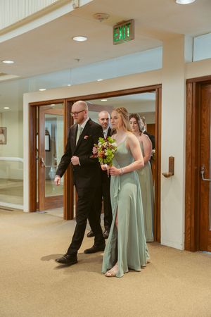 Wedding party members in mint green dresses and dark suits gather in a carpeted hallway before the ceremony.