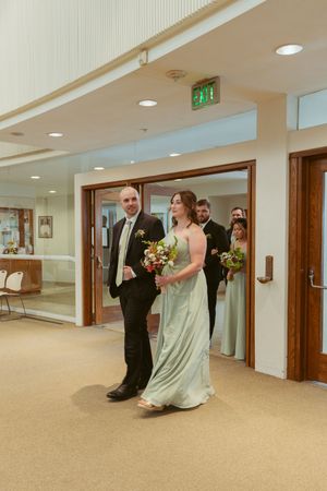 A couple walking down a carpeted hallway, the woman in a light dress holding a bouquet and man in dark suit.