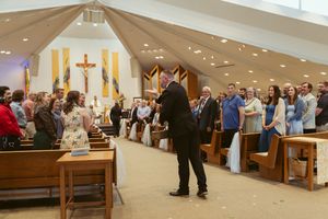 Wedding guests standing in church pews during ceremony with crucifix and organ pipes visible.