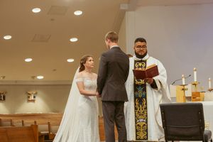 A wedding ceremony takes place at an indoor chapel with recessed lighting and an officiant at the altar.