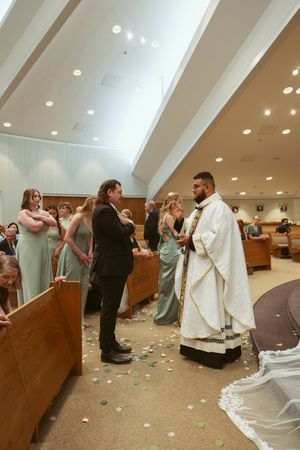 Inside a church, a religious ceremony takes place with attendants in formal attire and petal-scattered aisles.