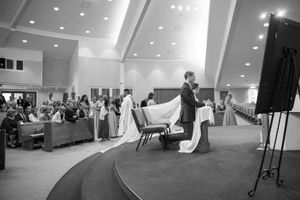A wedding veil trails along a church aisle during a ceremony ceremony with guests watching from the pews.