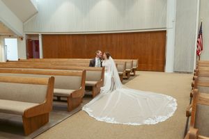 Long flowing white wedding veil stretches across wooden church pews in dramatic lighting.