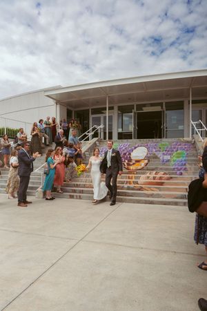 Wedding guests gather on colorful painted steps outside a modern building under cloudy skies.