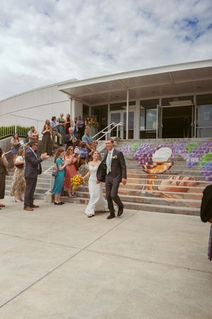 Wedding guests gather on steps outside a modern building with graffiti art under a cloudy sky.
