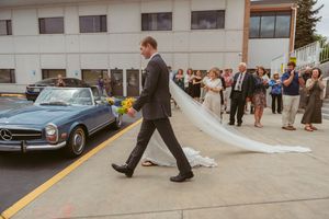 Wedding party and guests mingle outside church building near vintage car in parking area.