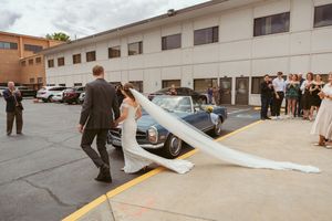 A couple walks through a parking lot with a long flowing train of white fabric trailing behind them.