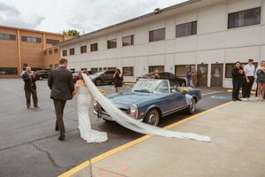 Wedding party gathers around classic blue convertible car in parking lot on cloudy day.