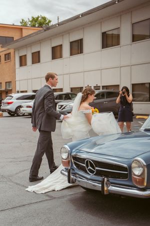 A classic Mercedes-Benz sits in a parking lot as wedding attendees in formal attire stand nearby.