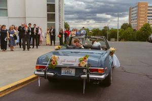 Group stands by decorated vintage blue car in lot with storm clouds overhead.