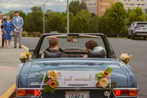 A vintage blue convertible decorated with flowers and 'just married' sign departing from a parking lot.