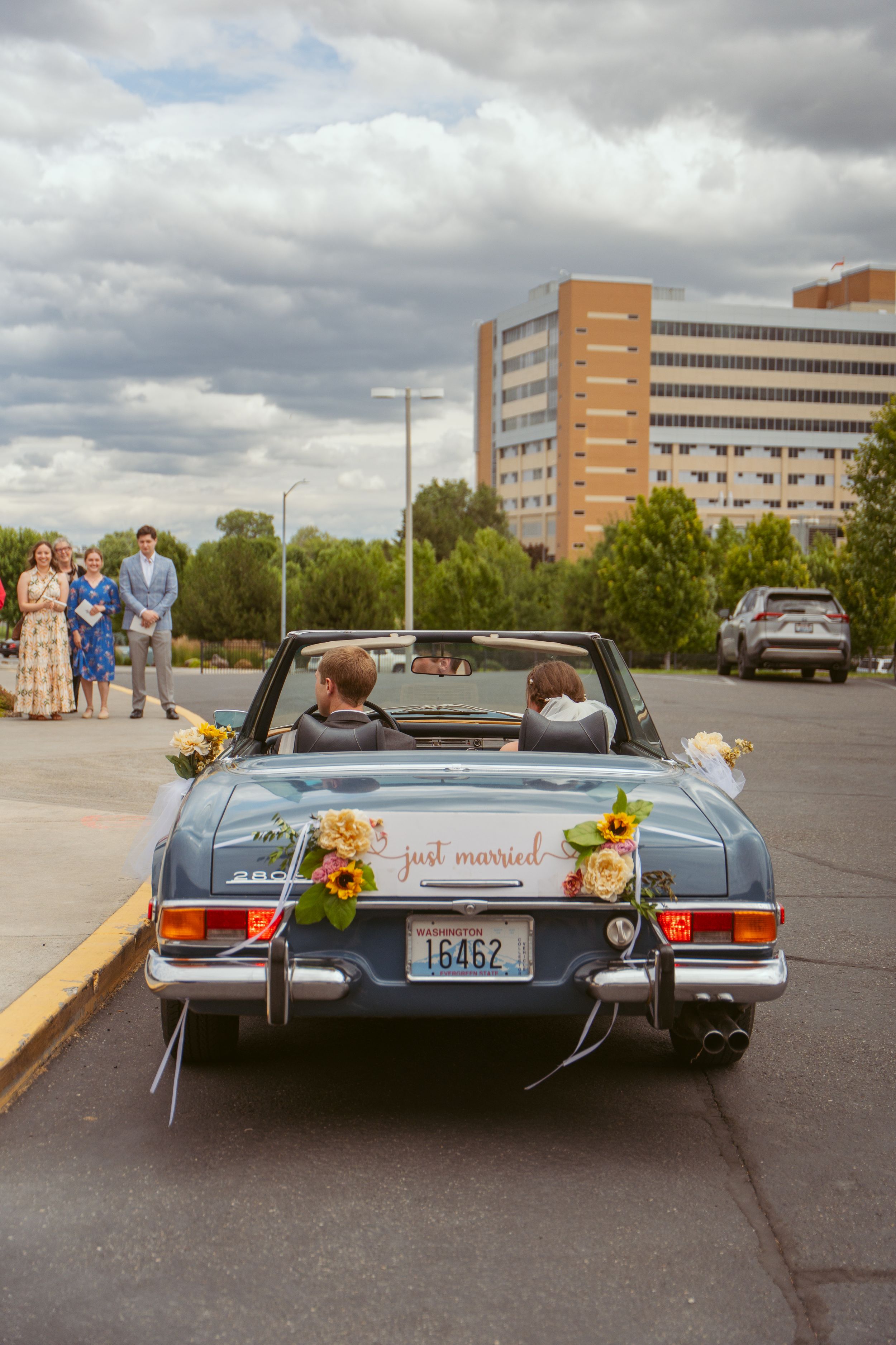 Classic convertible decorated with flowers and 'Just Married' sign parked in an empty lot with buildings in background.