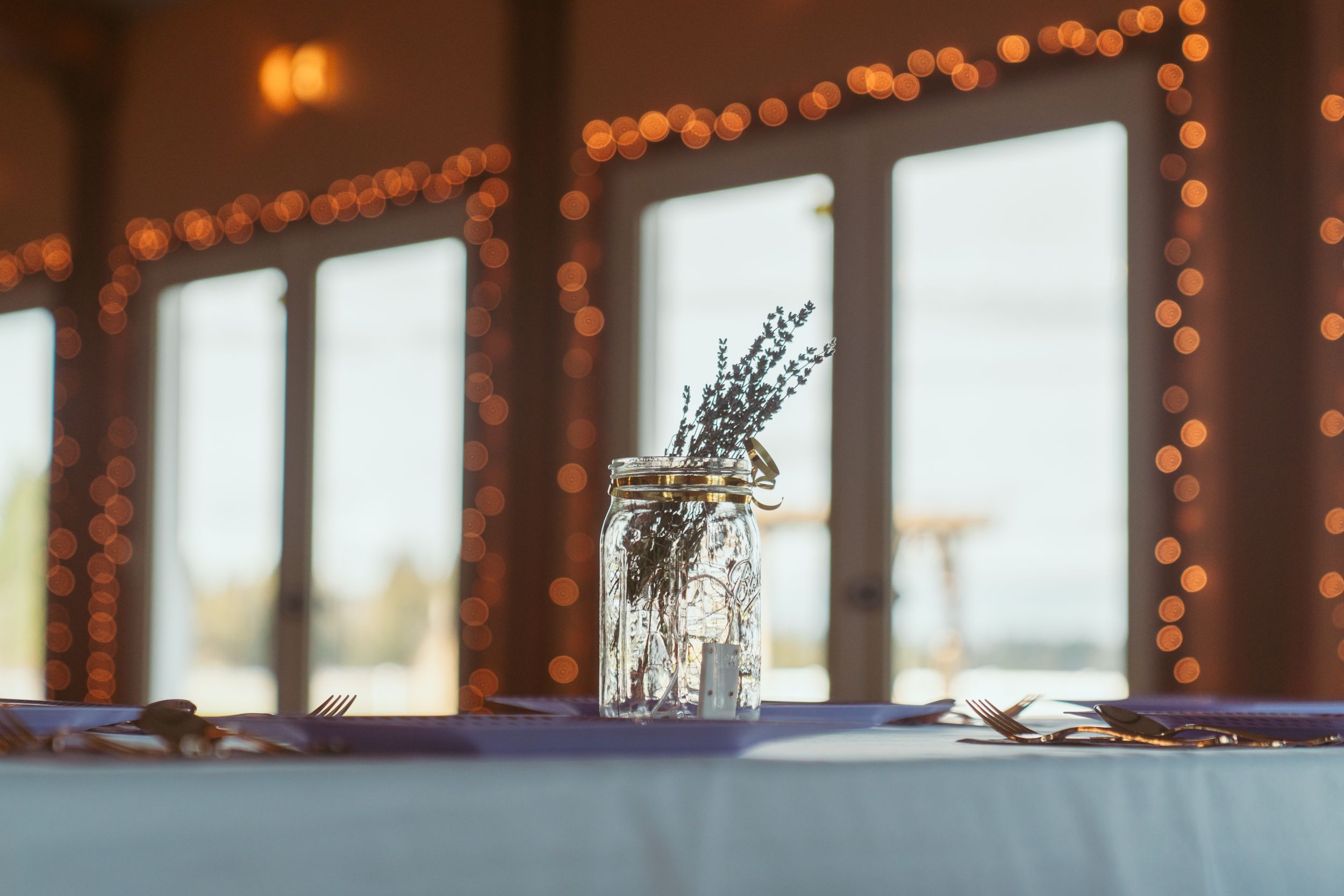 A frosted glass mason jar with dried flowers sits on a table near windows decorated with warm twinkle lights.