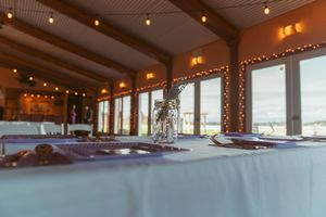 Catering buffet setup with place settings at a rustic venue featuring large windows and string lights along the ceiling.