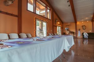 Empty event hall or dining room with white table linens, sloped wooden ceiling and windows.