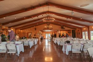 Indoor wedding reception venue with white table settings, string lights and wooden beam ceiling in warm lighting.
