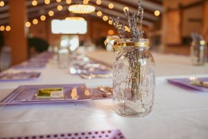 Mason jar centerpiece with lavender table settings at rustic wedding reception with string lights overhead.
