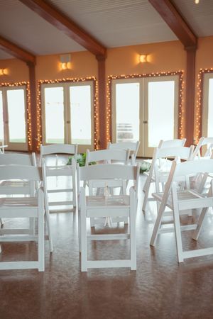 White wooden folding chairs line a bright indoor venue with string lights and exposed beams.