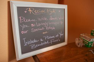 Chalkboard sign requesting guests to write in a recipe book displayed on wooden surface.