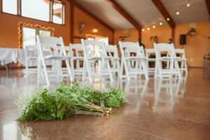 Row of white folding chairs set up for an indoor wedding ceremony with string lights overhead.