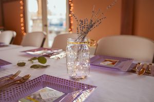 Mason jar with lavender flowers used as centerpiece on table with purple placemats and string lights in background.