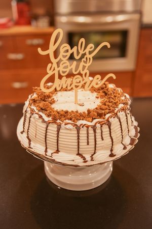 White frosted cake with chocolate drizzle and 'Love' topper on a cake stand.