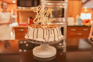 Elegant white layer cake with gold 'Love' topper and chocolate drizzle on display.