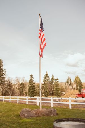 American flag flying on a pole beside white fencing on a grassy hillside with evergreen trees.