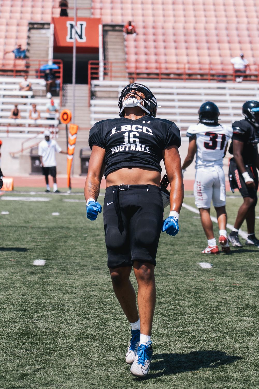 Football player walking on field wearing LBCC uniform with stadium seating in background.