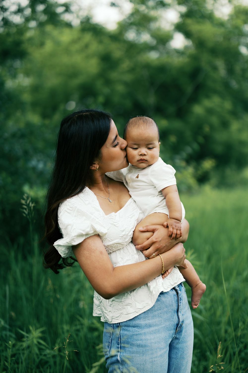 A tender moment between parent and baby in a white top and jeans standing against lush green foliage outdoors.