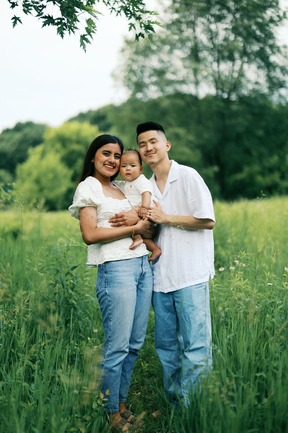 A couple holding their baby stands together in a lush green meadow on a summer day.