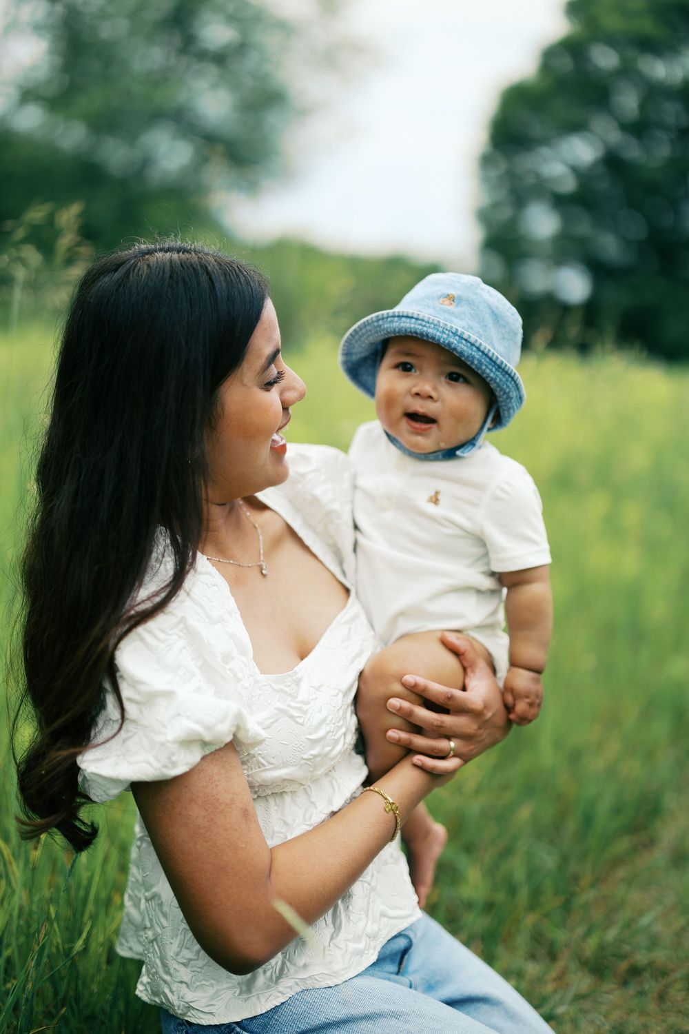 A woman in a white dress sits in a grassy field holding a baby wearing a blue knit hat on a sunny day.